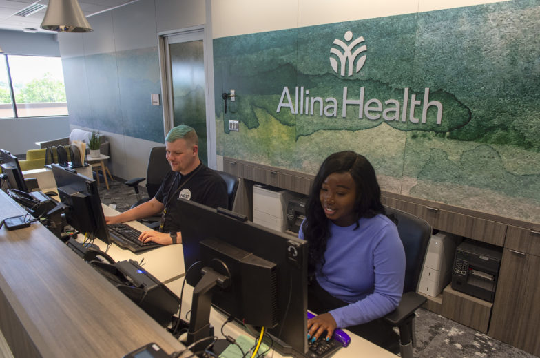 African american woman and white male sit and looking at monitors behind a reception desk at an Allina Health clinic